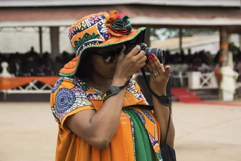 Une photographe à l'œuvre lors d'une cérémonie en plein air, en robe wax orange et verte.